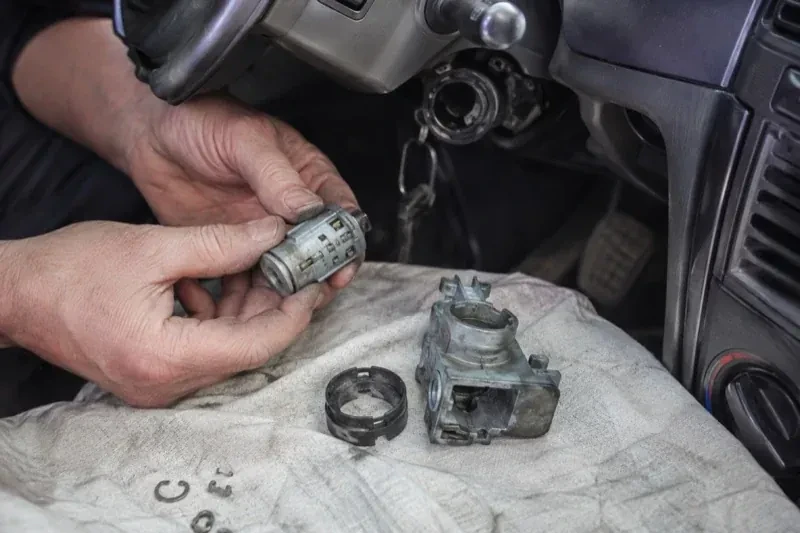 Hands holding a removed ignition cylinder beside a car steering column, with the housing and small parts on a cloth. — Ignition Lock Repair, Pickering